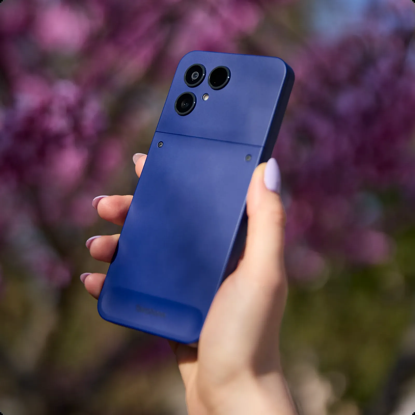 A hand holding a cobalt blue Fairphone 6 against a backdrop of pink spring blossom