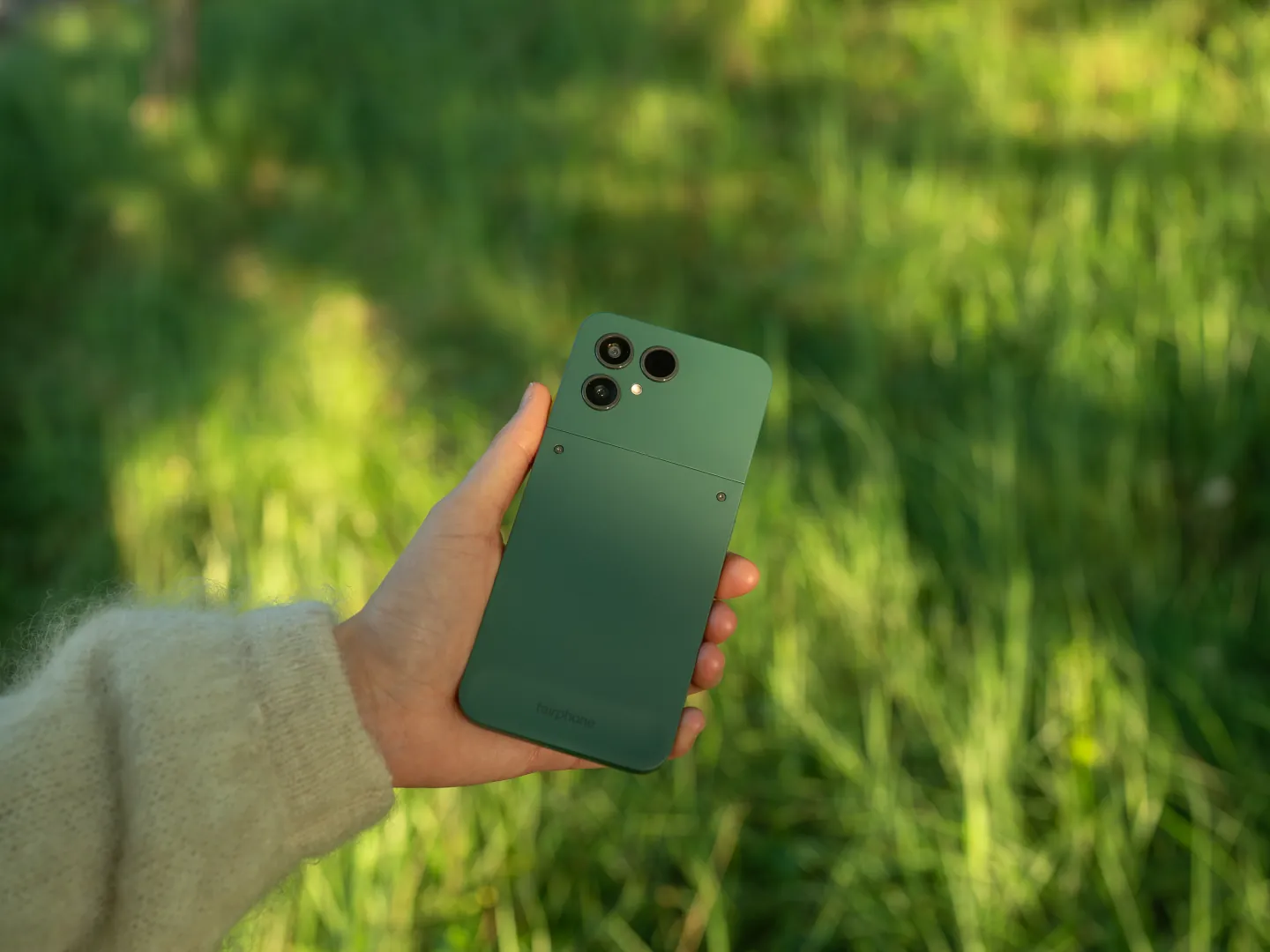 A hand holding a forest green Fairphone 6 above tall summer grass